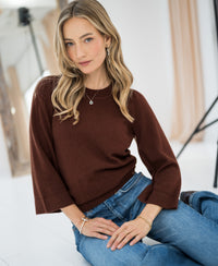 Woman wearing a cocoa brown Merino wool sweater by PLEIN PUBLIQUE, seated on the floor in a bright room.