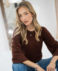 Woman wearing a brown merino wool sweater sits indoors, looking at the camera.