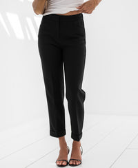 Black 7/8-length Faubourg pants shown on model, standing on white floor with torso cropped.