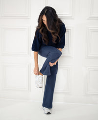 Woman wearing a PLEIN PUBLIQUE LA FRAMBOISE dark blue sweater stands by a white paneled wall.