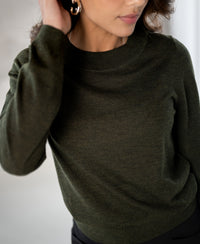 Woman wears the PLEIN PUBLIQUE merino wool sweater LA GIGI in bottle green, blurred background.