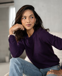 Woman wearing dark violet PLEIN PUBLIQUE Merino wool sweater LA GIGI, seated on a couch, looking to the side.