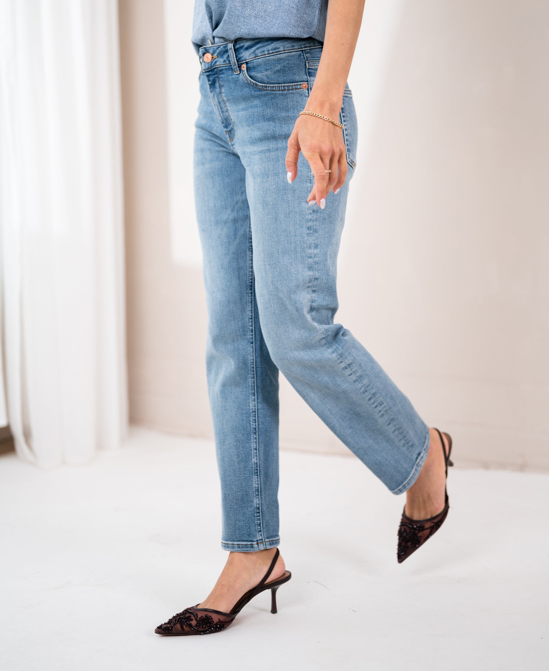 Model wears light stone-wash cropped jeans, standing indoors with one leg raised.