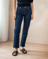 Person wearing cropped jeans in a dark rinse wash, standing on light wood floor near a white curtain.