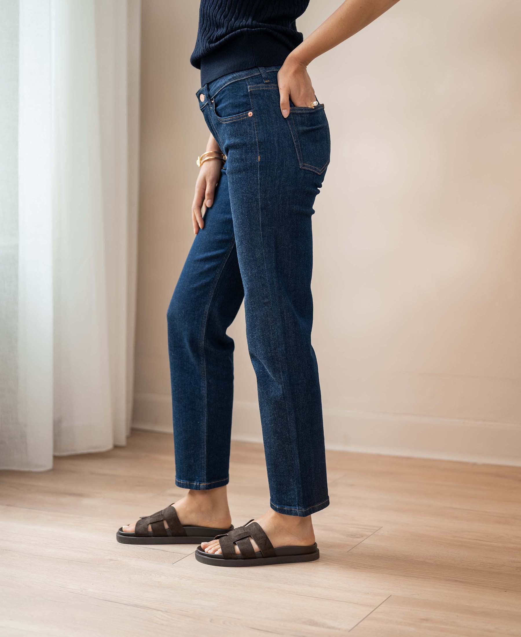 Person wears blue cropped jeans in a rinse wash from PLEIN PUBLIQUE, standing by a white curtain.