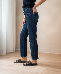 Person wears blue cropped jeans in a rinse wash from PLEIN PUBLIQUE, standing by a white curtain.