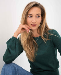 Woman wearing greenish PLEIN PUBLIQUE LA COEUR merino wool sweater, sitting and looking to the side.