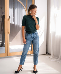 Woman wearing a PLEIN PUBLIQUE dark green merino wool sweater stands indoors by sunlight, looking down.
