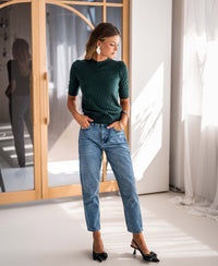 Woman wearing a dark green PLEIN PUBLIQUE merino wool sweater, standing in a bright room.
