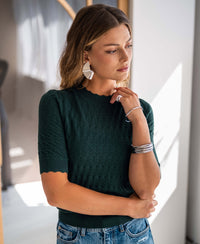 Woman wears a dark green PLEIN PUBLIQUE Merino wool sweater LA BRIOCHE, standing indoors.