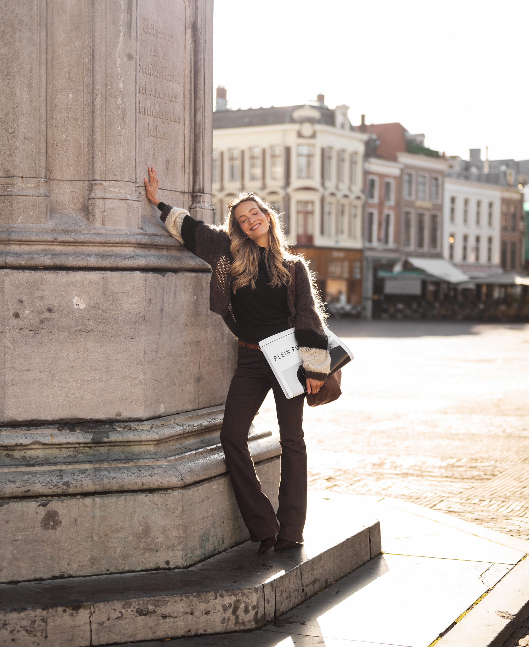 Woman wearing a brown PLEIN PUBLIQUE fluffy vest stands by a pillar.