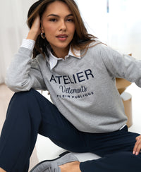 Woman indoors wears light grey LA HARPER sweater by PLEIN PUBLIQUE, looking at the camera.