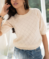 Woman wears the Argyle merino wool sweater in butter yellow from PLEIN PUBLIQUE in a bright room.