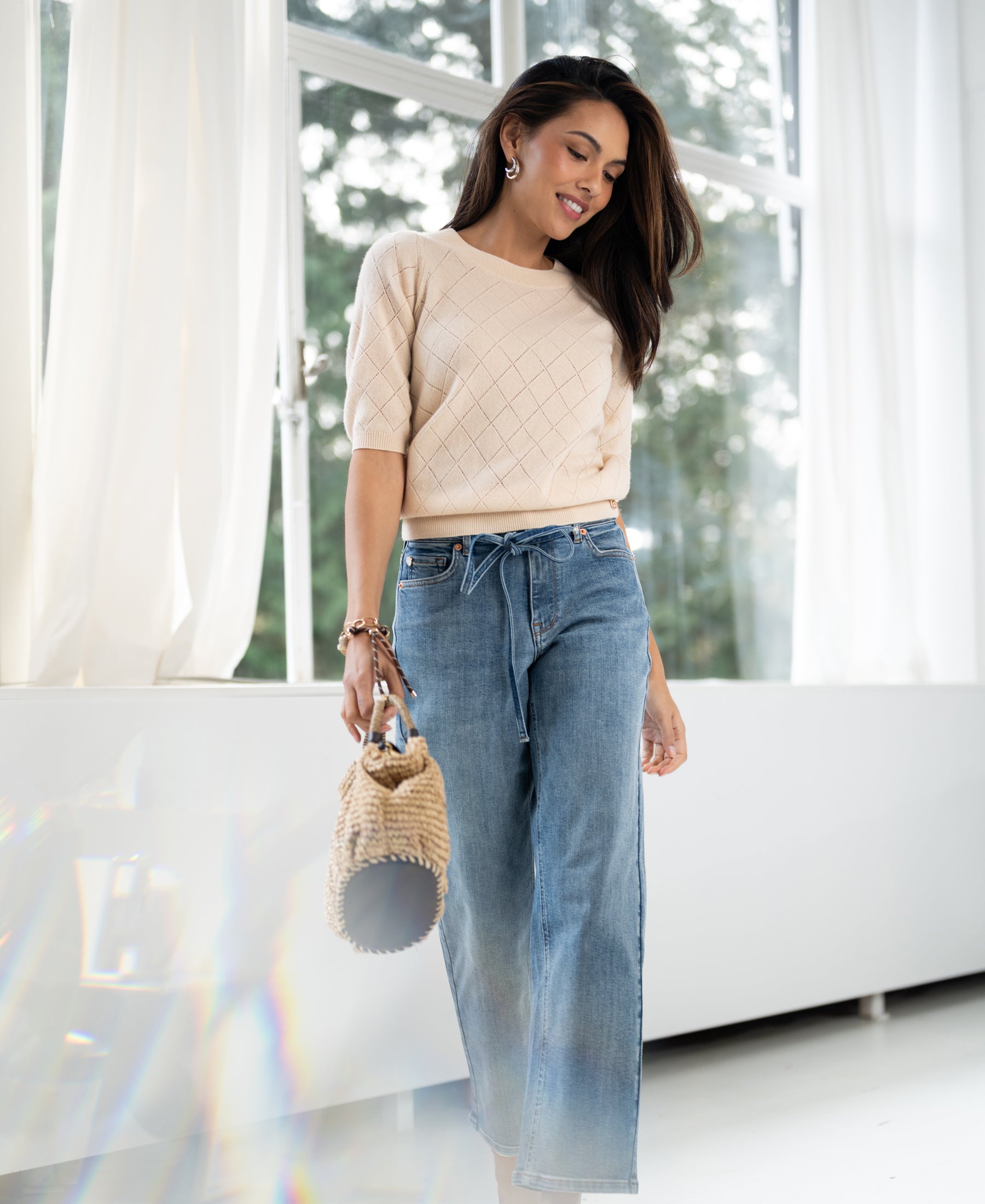 Light stone cropped jeans, worn by a woman standing by curtained windows.