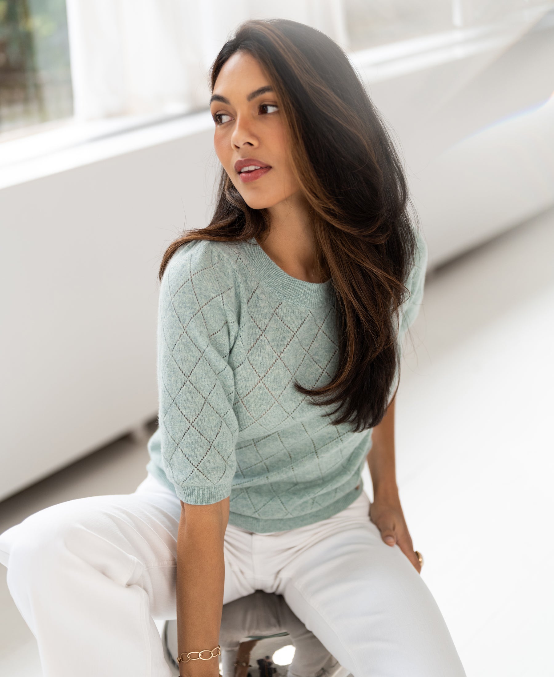 Argyle merino wool sweater in mist green, shown worn by a woman with long brown hair, seated in a bright room.