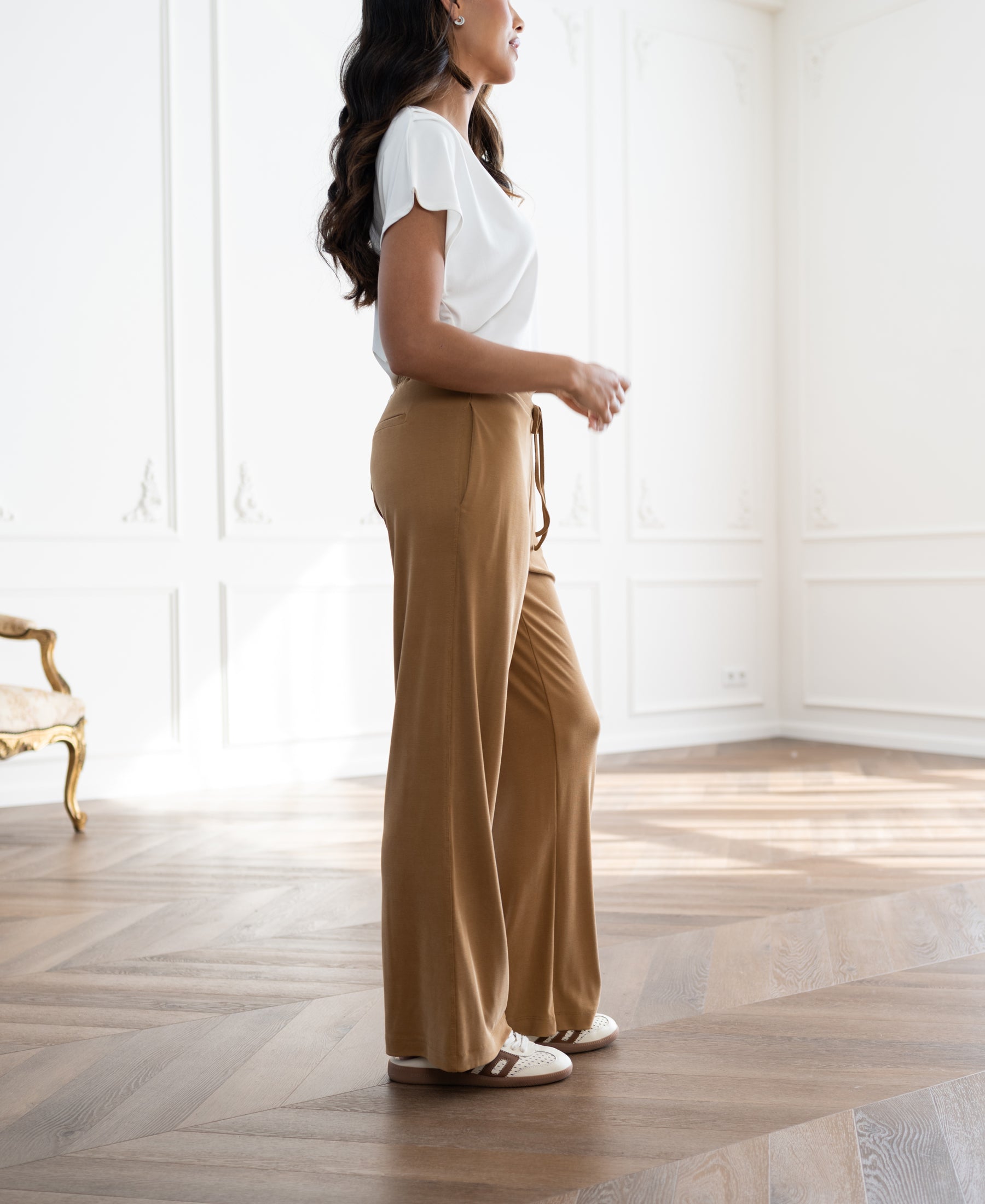 Light camel lounge pants by PLEIN PUBLIQUE, shown worn by a woman standing in a bright room.