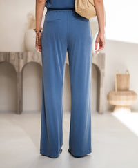 Blue lounge pants in soft fabric, shown indoors near a wooden table and wicker décor.
