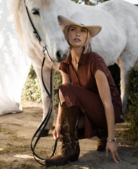 Woman wearing a camel-colored travel polo dress kneels by a white horse, holding its reins.
