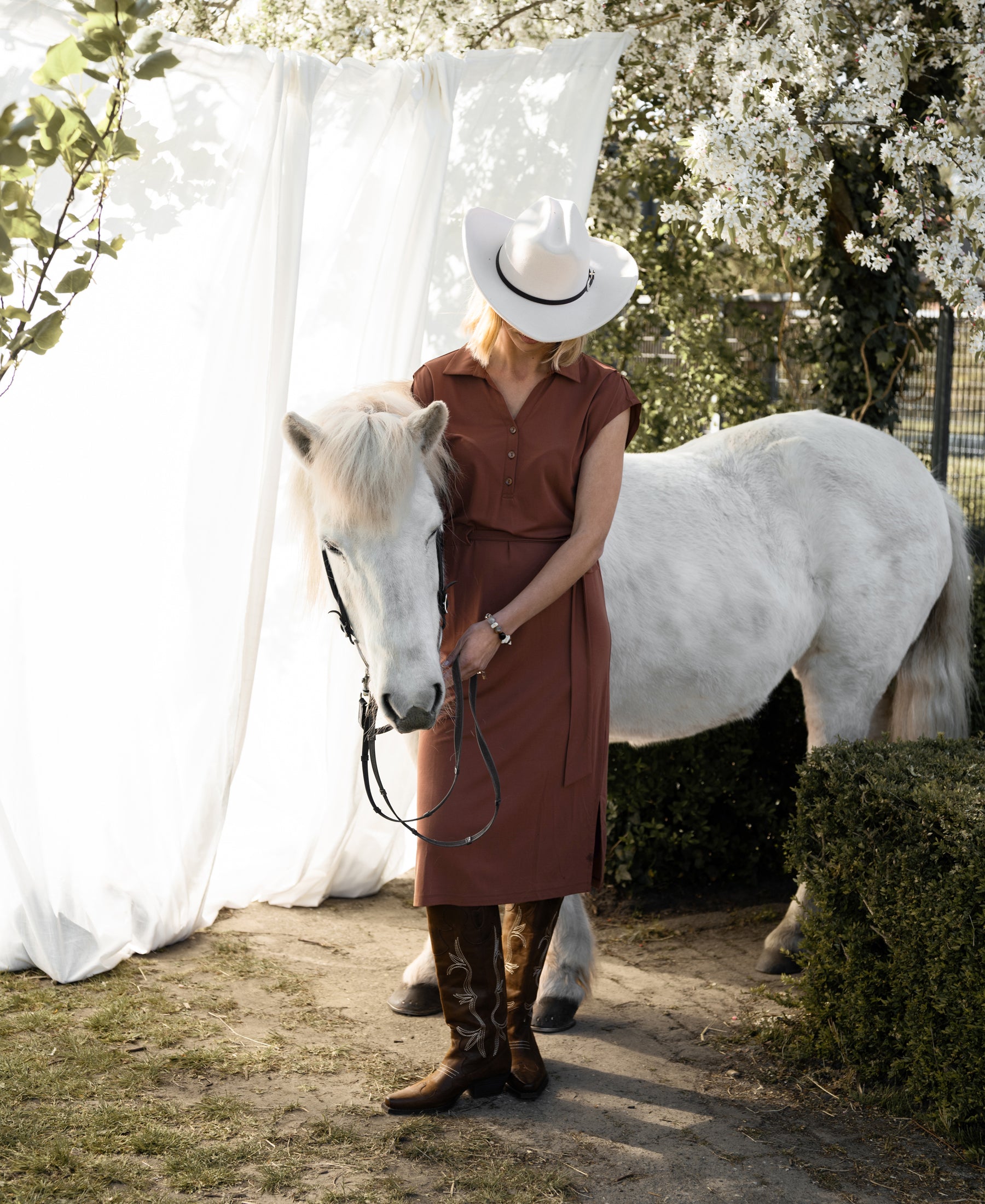 Woman wearing a camel-colored INES travel polo dress by PLEIN PUBLIQUE stands in a sunlit garden.
