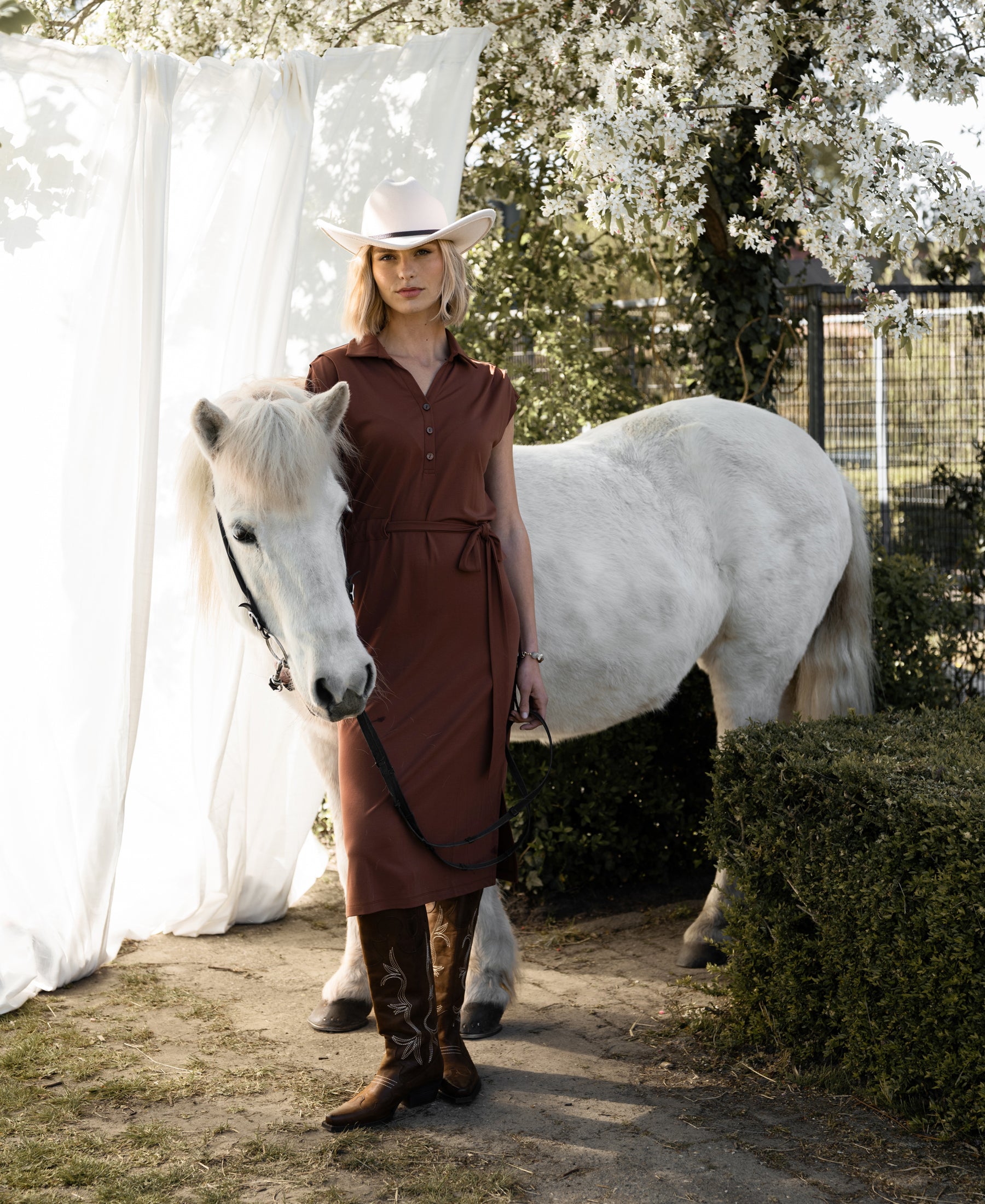 Woman wearing a camel PLEIN PUBLIQUE Travel polo dress INES stands beside a white horse, flowers in the background.