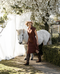 A woman wears a PLEIN PUBLIQUE Travel polo dress INES in camel under blooming trees in bright sunlight.