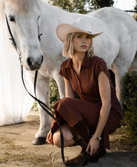 Camel-colored polo dress with travel-friendly fabric, shown on a model crouching by a white horse and holding its reins.