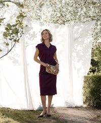 Woman wearing a wine-colored travel polo dress poses outdoors by white curtains.