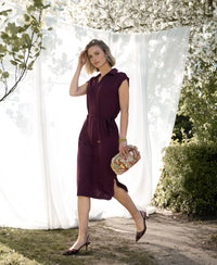 Woman wearing a wine-colored Ines Travel polo dress by Plein Publique, standing outdoors with trees in the background.