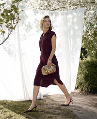 Woman wearing a PLEIN PUBLIQUE Travel polo dress INES in wine color, white curtains and blooming trees in the background.