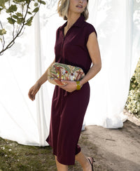 Woman wearing a wine-colored INES travel polo dress by PLEIN PUBLIQUE walks outdoors near greenery.