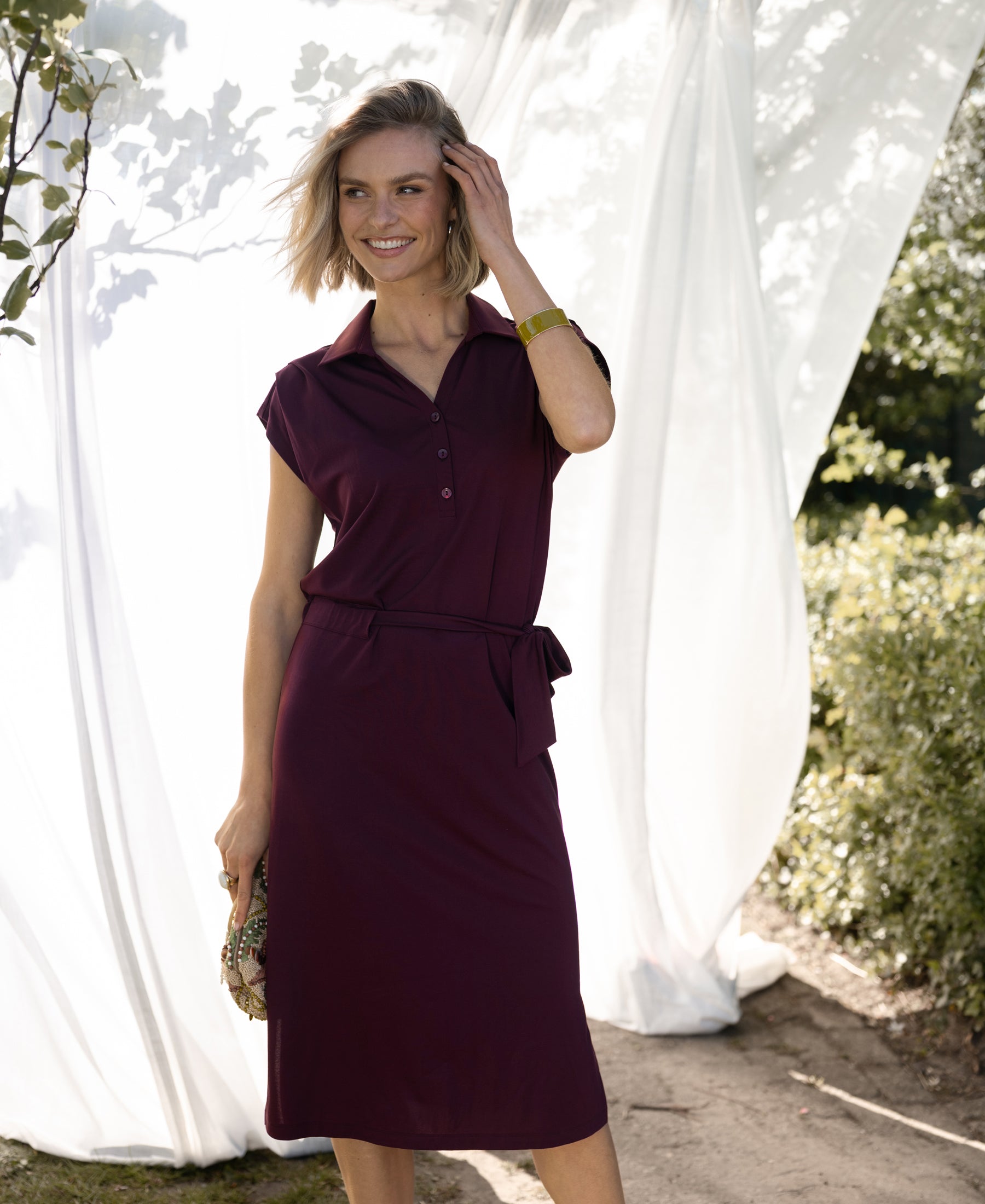 Woman wearing a wine-colored travel polo dress stands outside, white fabric backdrop behind her.