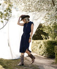 Woman wearing a PLEIN PUBLIQUE INES dark blue travel polo dress walks outdoors by white blossoms.