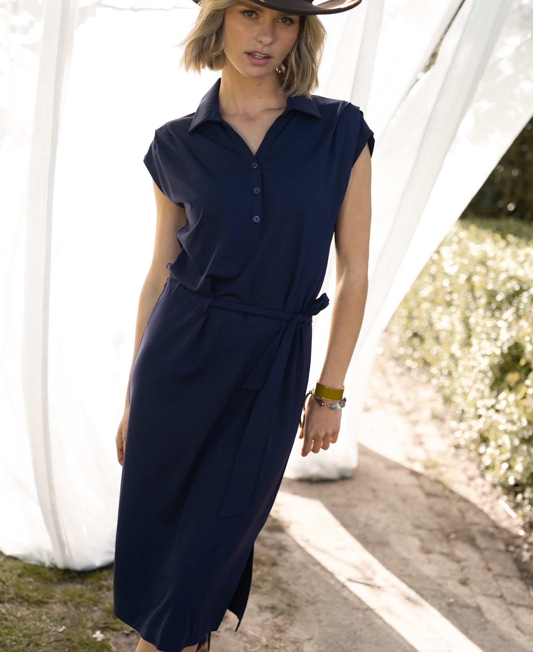 Navy blue PLEIN PUBLIQUE Travel polo dress INES shown outdoors near white curtains and greenery.