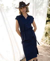 Woman wearing dark blue INES Travel polo dress by PLEIN PUBLIQUE, standing near white curtains in sunlight.