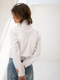 Back view of a woman wearing a white PLEIN PUBLIQUE denim jacket in a minimal room.