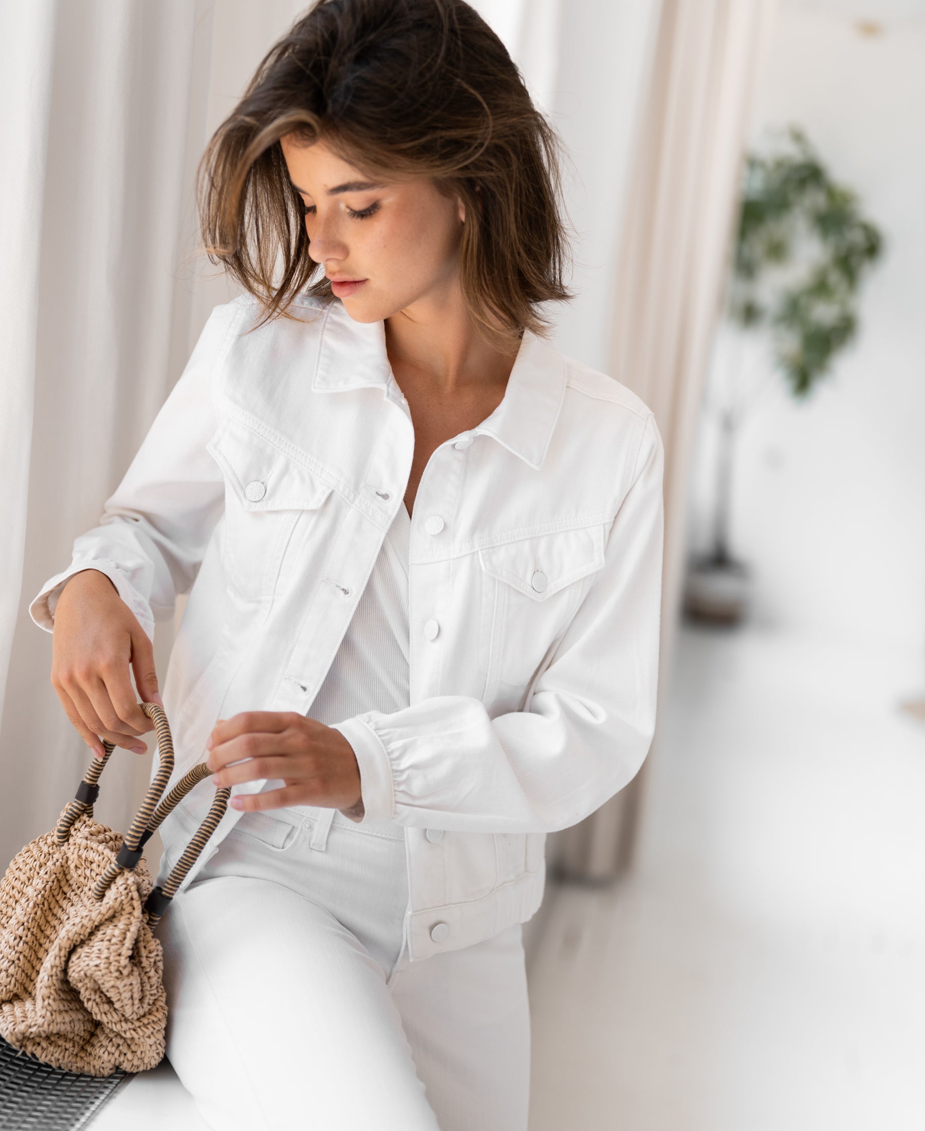 White denim jacket displayed by white curtains and a green plant.