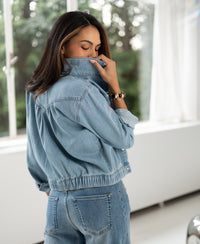 Back view of woman wearing a light-wash denim jacket indoors, lifting the collar and facing sunlight.