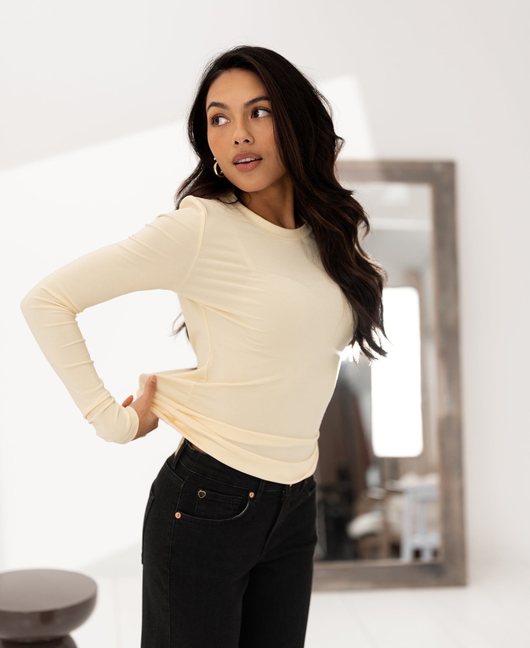 Woman wears PLEIN PUBLIQUE Luxe long-sleeve LA JANE T-shirt in soft yellow, standing indoors by a mirror.