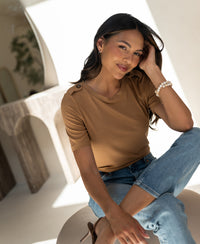 Woman wearing a yellow bamboo T-shirt sits indoors, smiling and touching her hair.