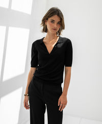 Woman wears black PLEIN PUBLIQUE Tencel-silk polo shirt in a bright room with white walls and window shadows.