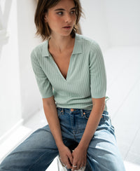 PLEIN PUBLIQUE’s Tencel-silk polo shirt in light green seen on a woman seated in a bright, minimal room.