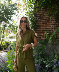 Woman wearing olive green LA JODY blouse by OVERIG stands outside by a brick wall with flowers on a sunny day.