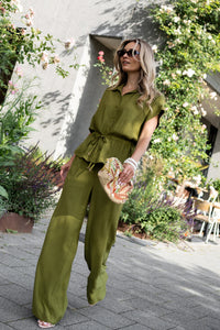 Woman wearing olive green LA LYNETTE pants by OVERIG stands outdoors near greenery.