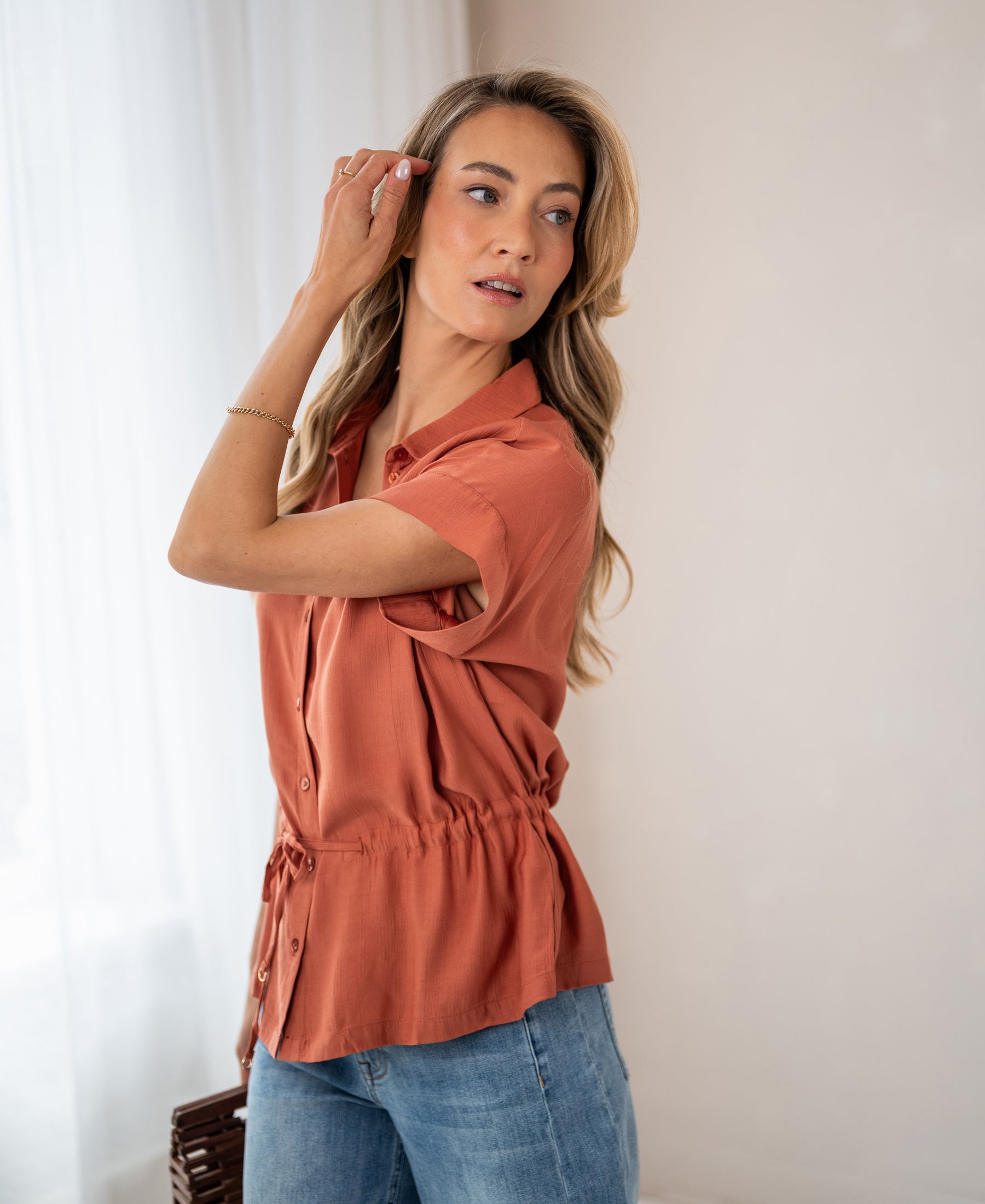 Woman wears the LA JODY Terra blouse in a terra cotta color by OVERIG, standing near a window.