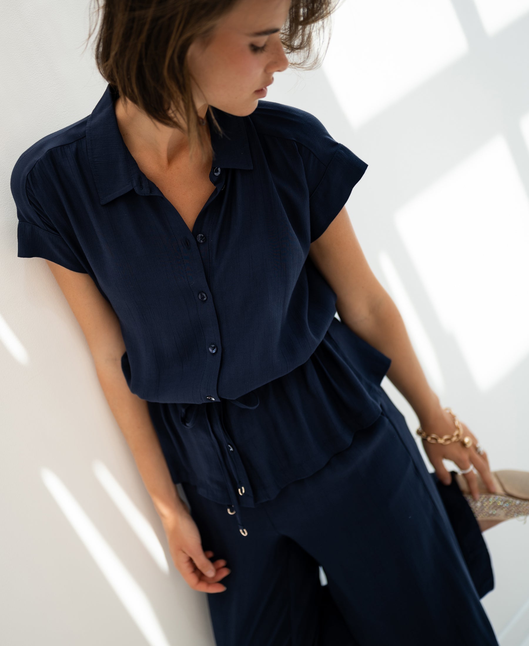 Woman wearing a dark blue LA JODY blouse stands by a sunlit white wall.