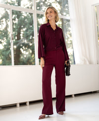 Woman wearing burgundy LA JOELLE trousers by PLEIN PUBLIQUE, standing by large windows.