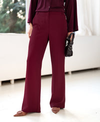 Person wearing soft LA JOELLE wine-colored pants by PLEIN PUBLIQUE, standing by a window.