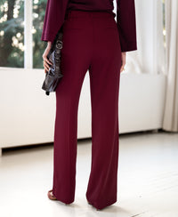 Woman wearing LA JOELLE Wine Smooth Pants from PLEIN PUBLIQUE, standing by large windows with white curtains.