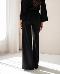 Person standing indoors in sunlight, facing away, wearing black PLEIN PUBLIQUE LA JULIA travel pants.