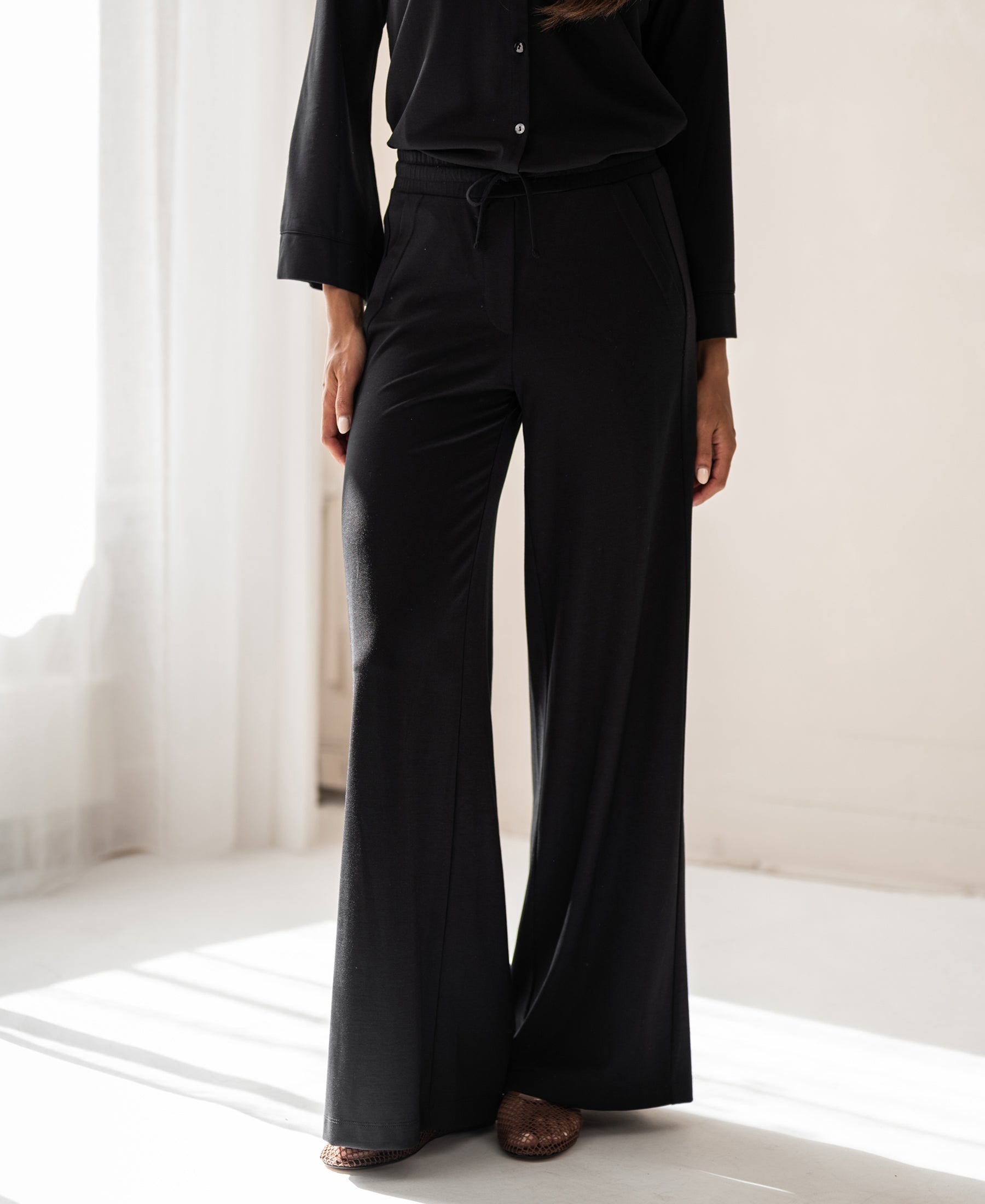 Woman wears black PLEIN PUBLIQUE LA JULIA travel pants indoors in natural sunlight; wrinkle-resistant quality.